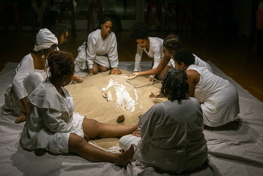 Group_Of_Black_Performers_Wearing_White_Clothing_Scooping_Sand_Onto_A_Woman_Lying_On_The_Floor