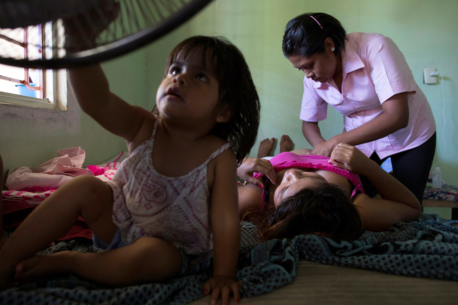 Midwife_Pressing_Belly_Of_Pregnant_Mexican_Woman_Lying_Down_With_Young_Child_In_Foreground