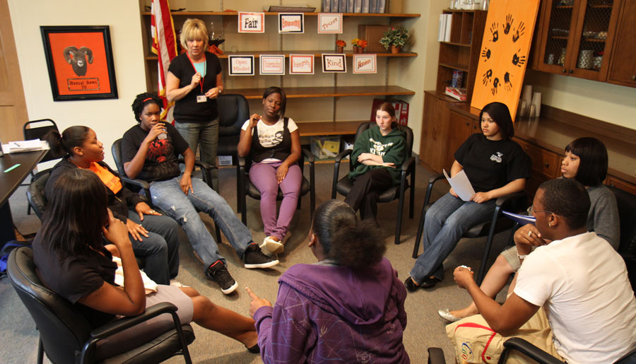 Teenagers Sitting In Circle With Facilitator Standing And Leading The Group
