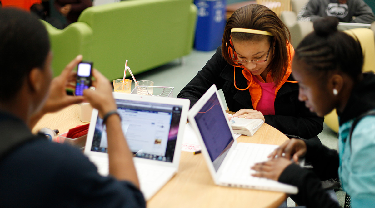 Three teen girls sitting at a table using computers and phone