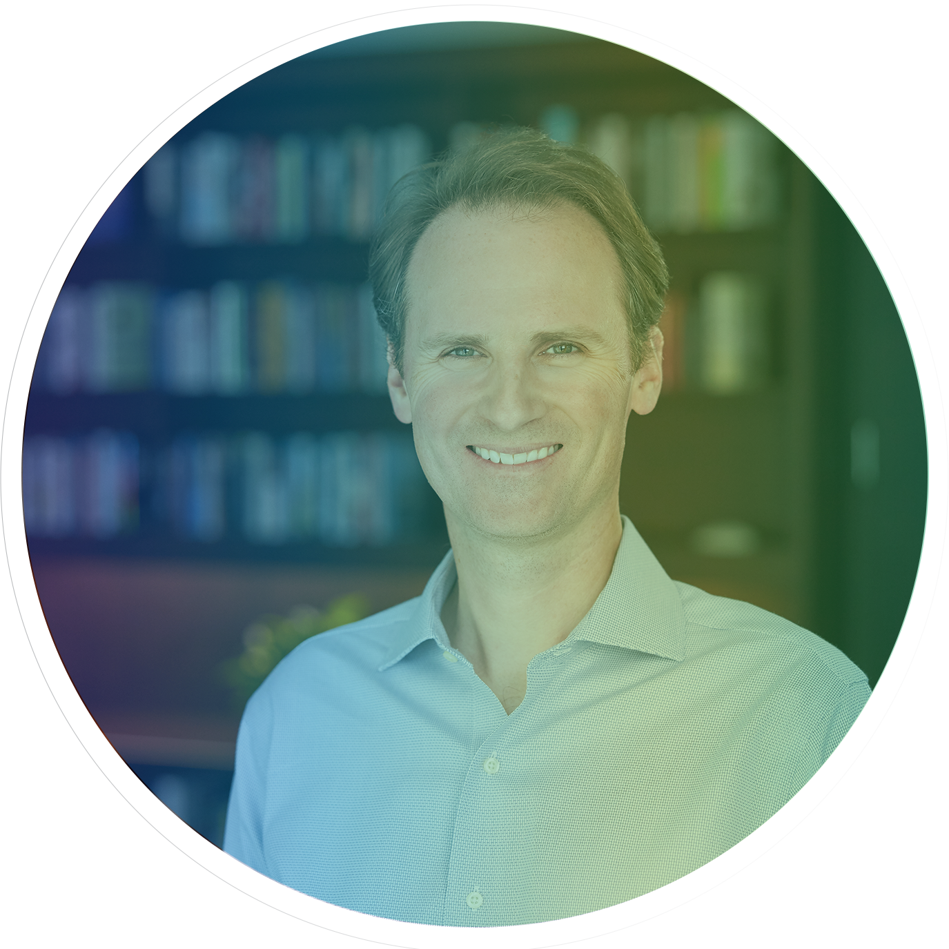 A smiling middle aged white man in a light blue button-down shirt stands in front of a book case